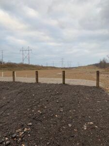 A gravel lot with a wooden fence line bordering an open, cleared field under a cloudy sky with distant power lines.