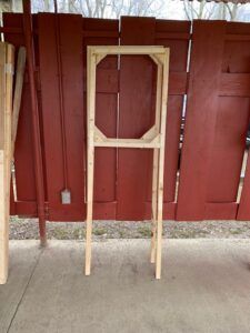 A tall, wooden rectangular frame structure with corner supports, standing outdoors against a red wooden fence.