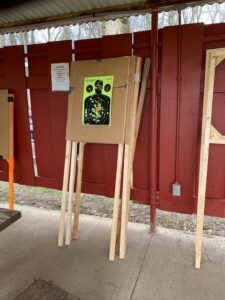 A target stand with a human-silhouette shooting target attached, set up against a red wooden wall at a firing range.