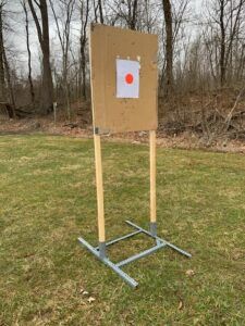 A target stand made of wood and metal holds a cardboard square with a paper target featuring a red bullseye in a field.