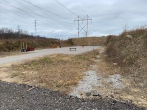 An outdoor gravel lot with a small wooden bench and metal structures, set beneath large high-voltage power lines.