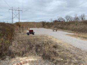 A gravel shooting range with a small wagon, target stands, and a bench, set against an earthen berm and power lines.
