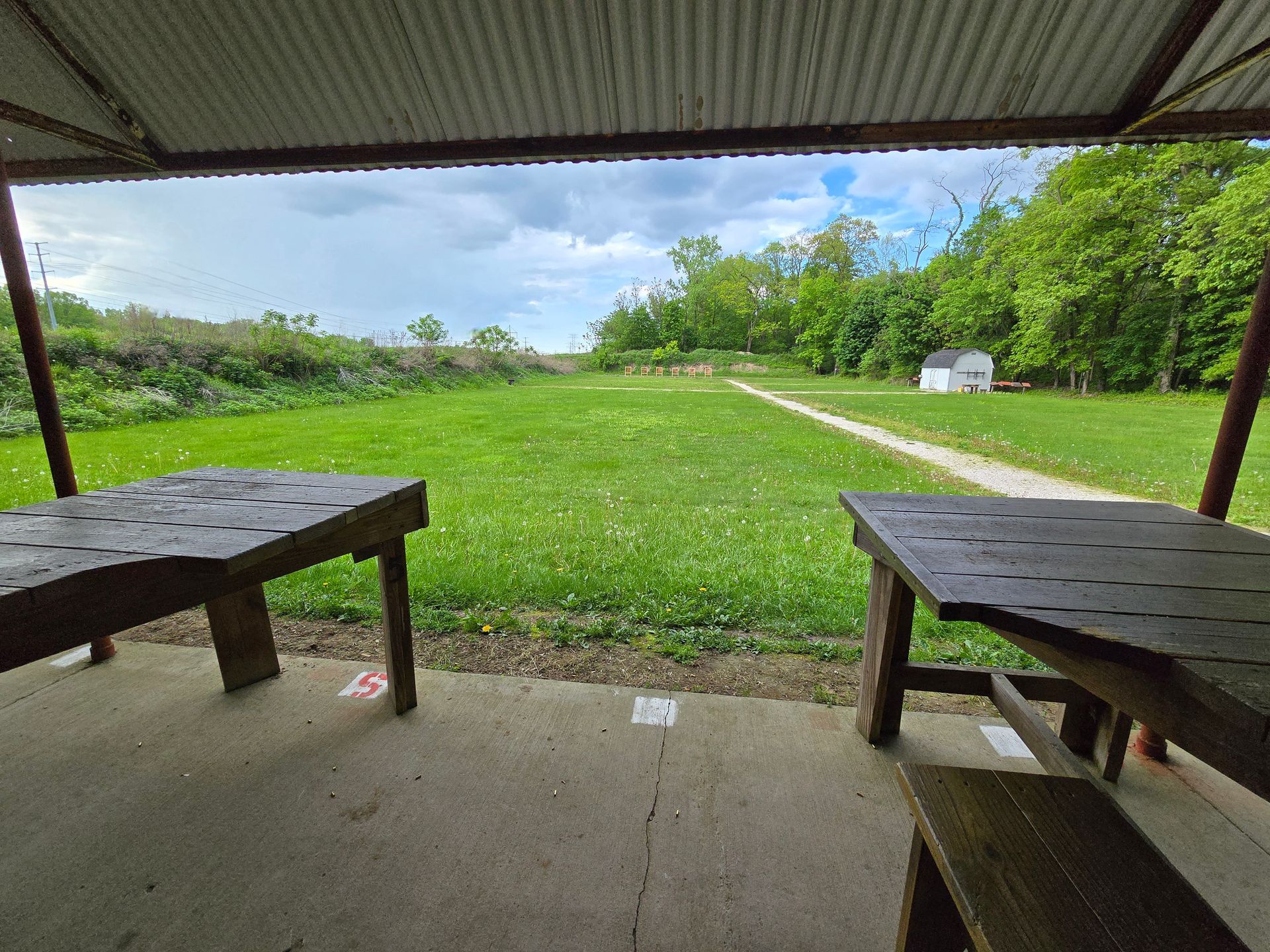 A covered outdoor shooting range with wooden benches, a grass field, and a white storage shed in the distance.
