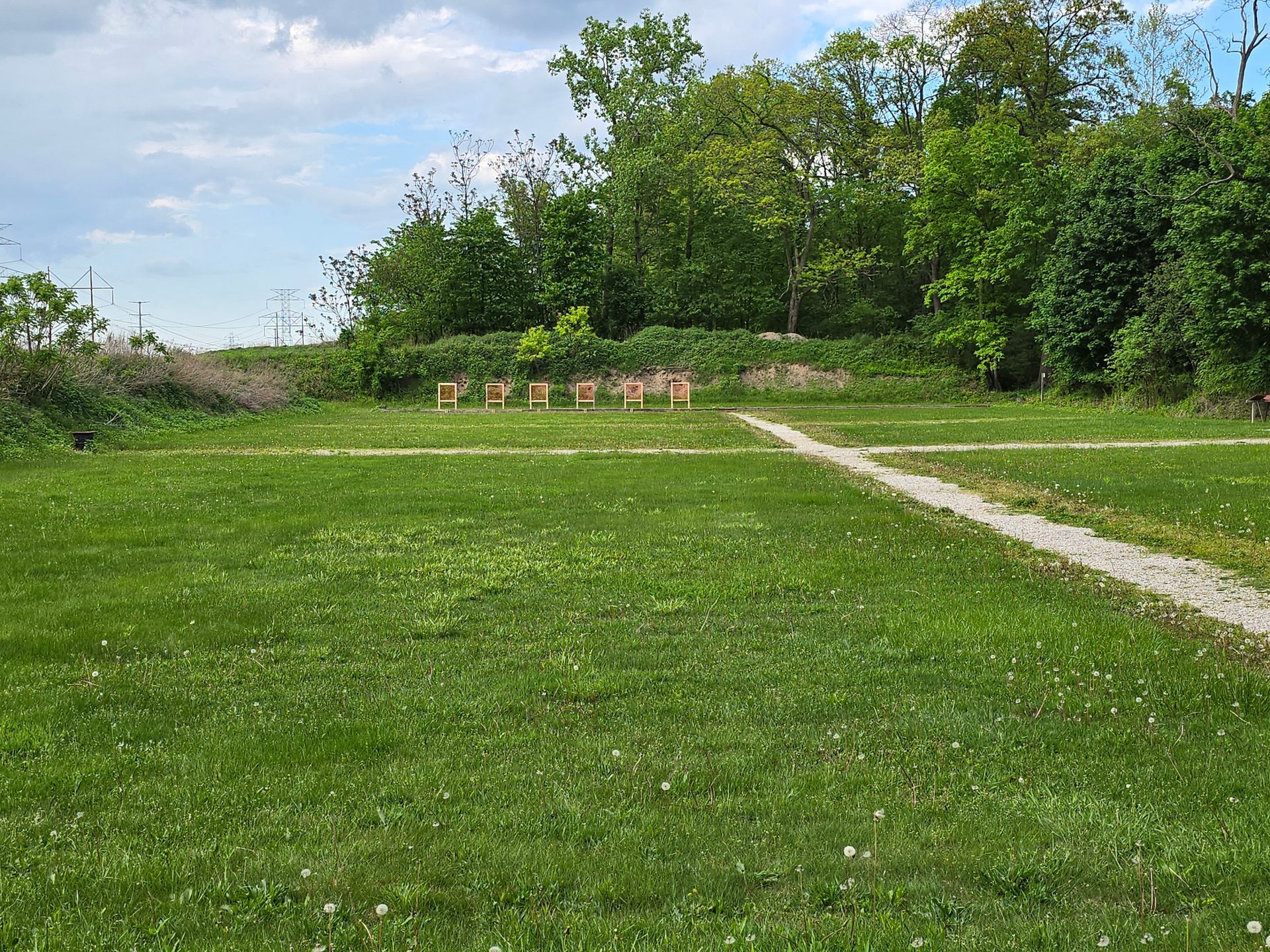 A grassy field with a gravel path and a row of targets in the distance, framed by trees under a partly cloudy sky.