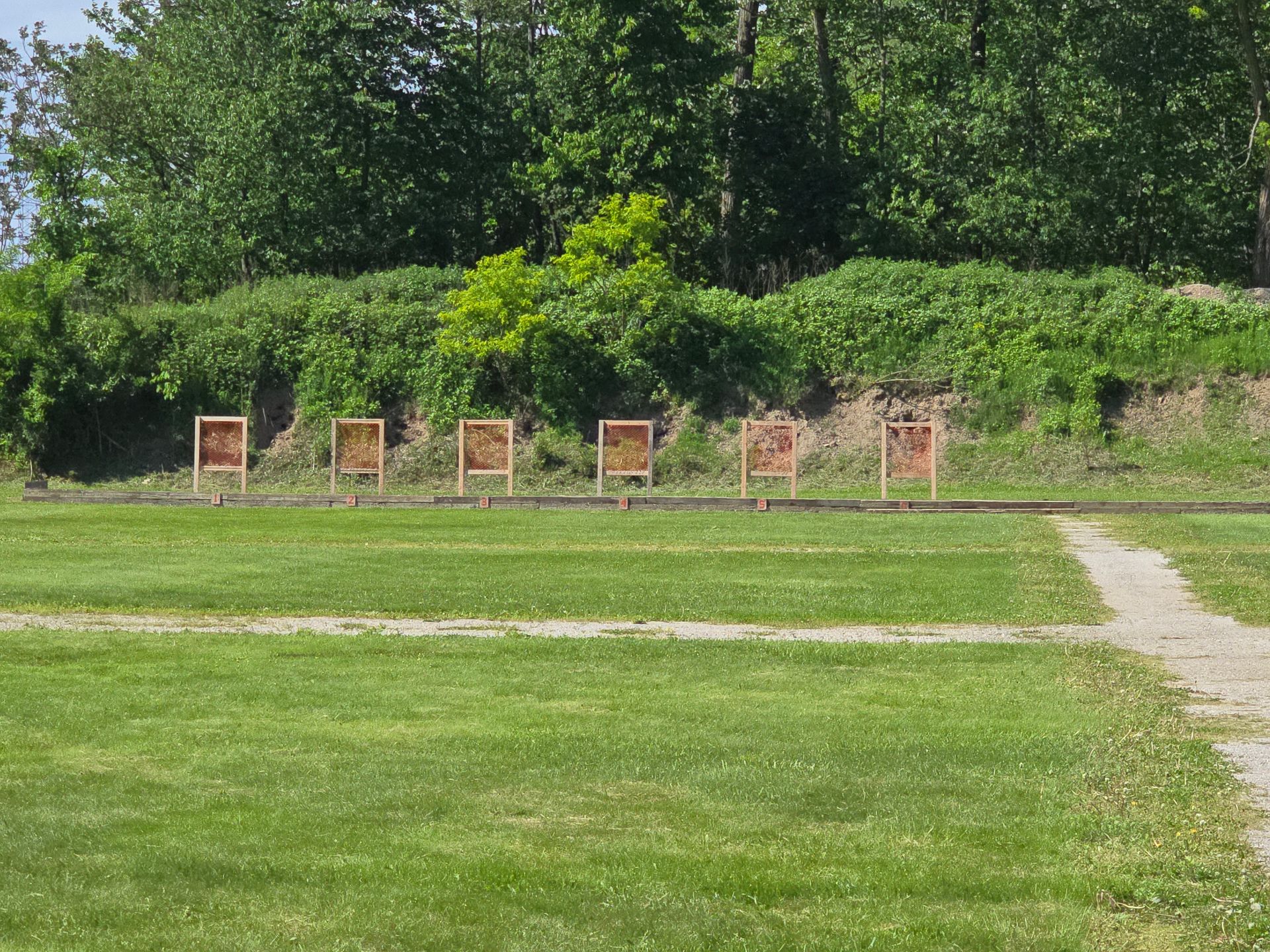 Six target stands arranged in a row in a grassy field in front of a wooded dirt embankment.