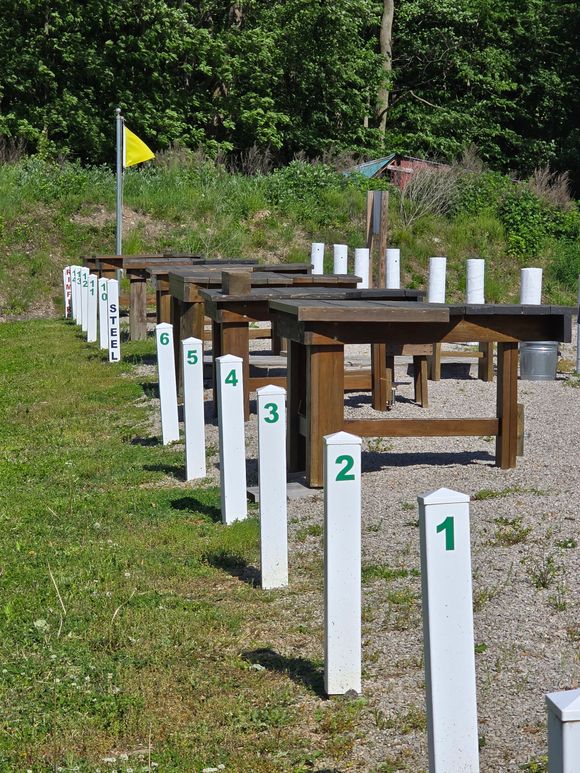 A line of numbered shooting range stations with wooden tables and white markers on a gravel path near green trees.