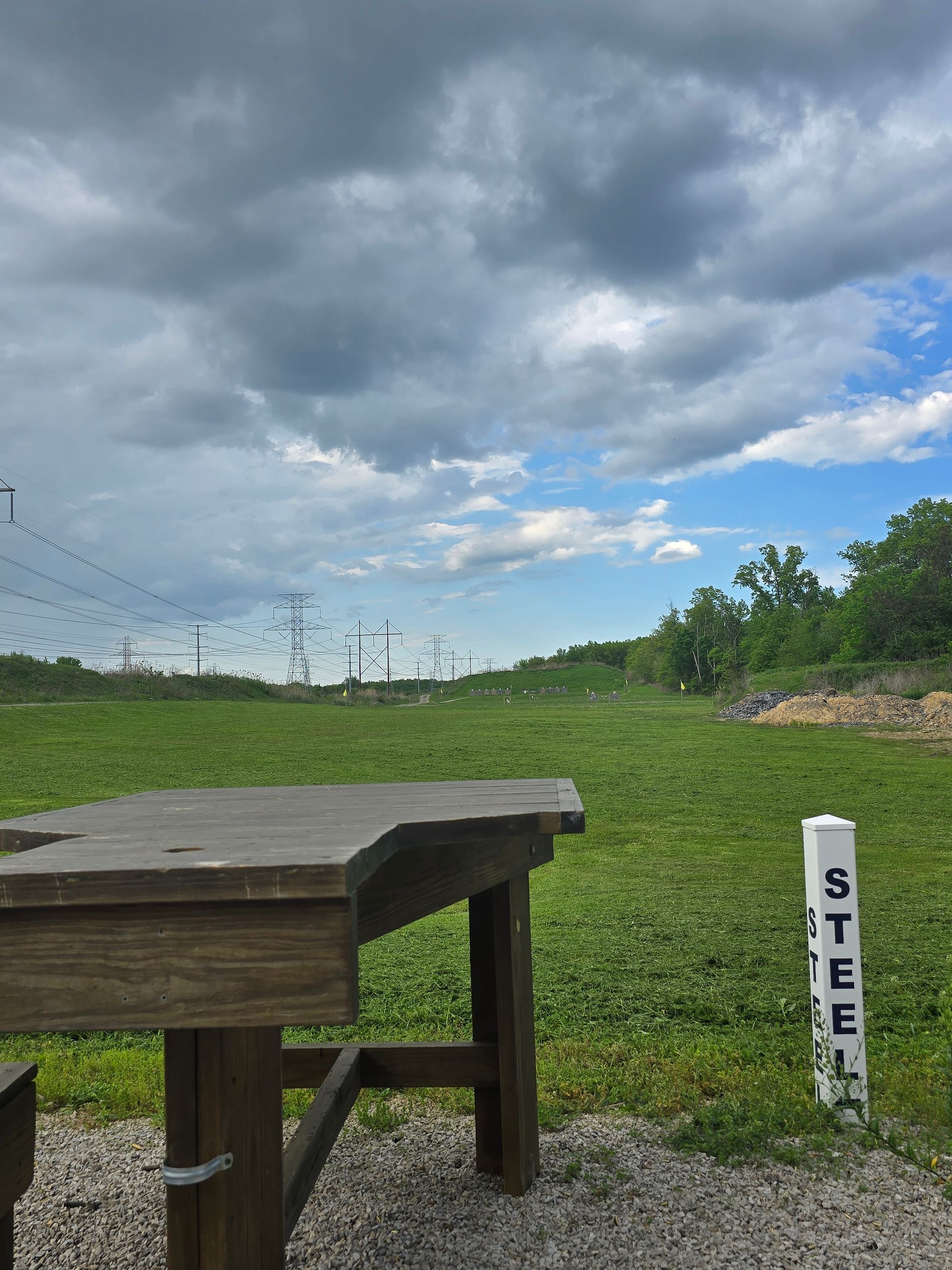 A wooden shooting bench sits on a gravel pad facing a grassy field under a cloudy blue sky, with a white