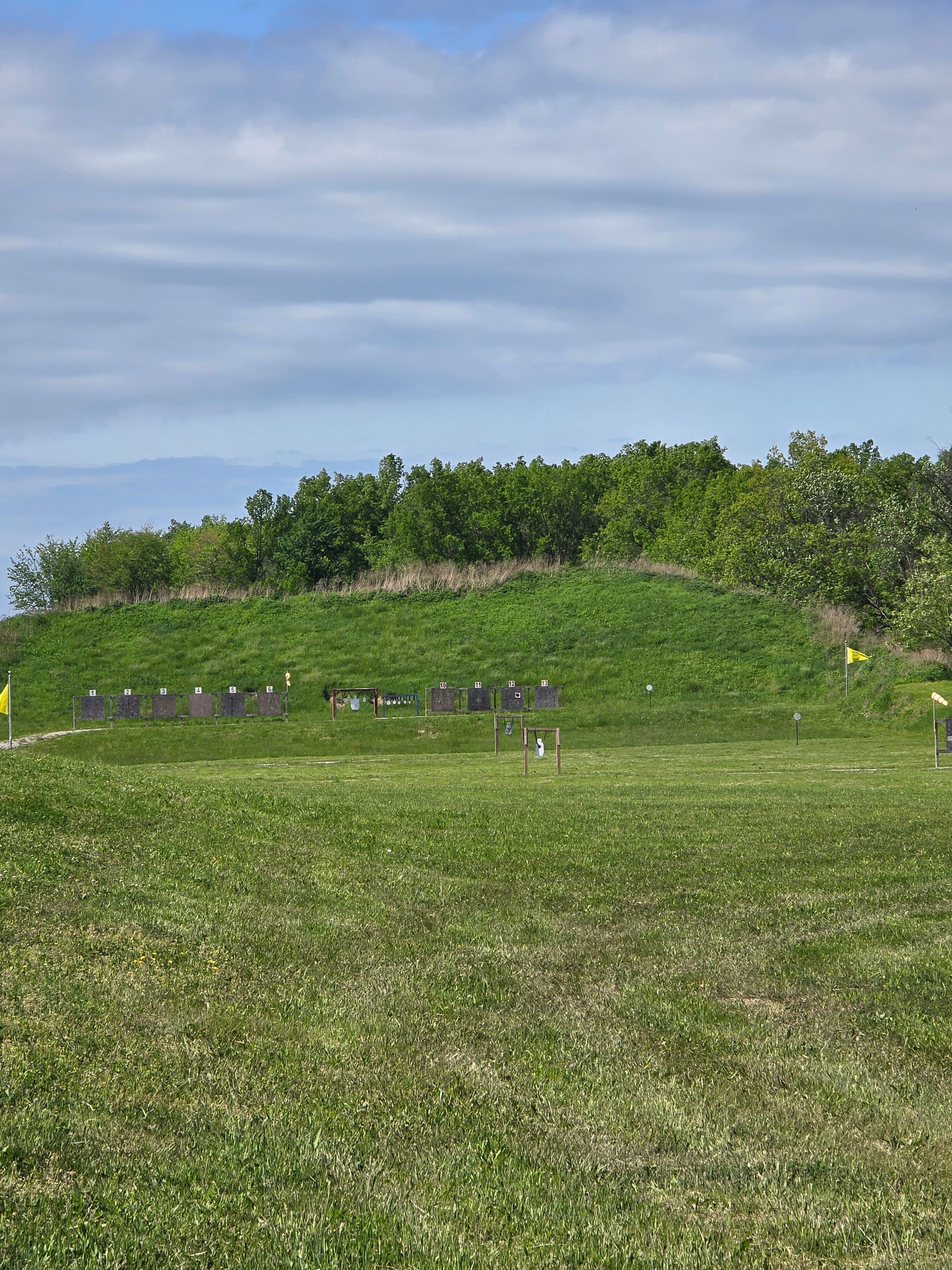 A green, grassy shooting range with target stands set against a low hill lined with trees under a cloudy blue sky.