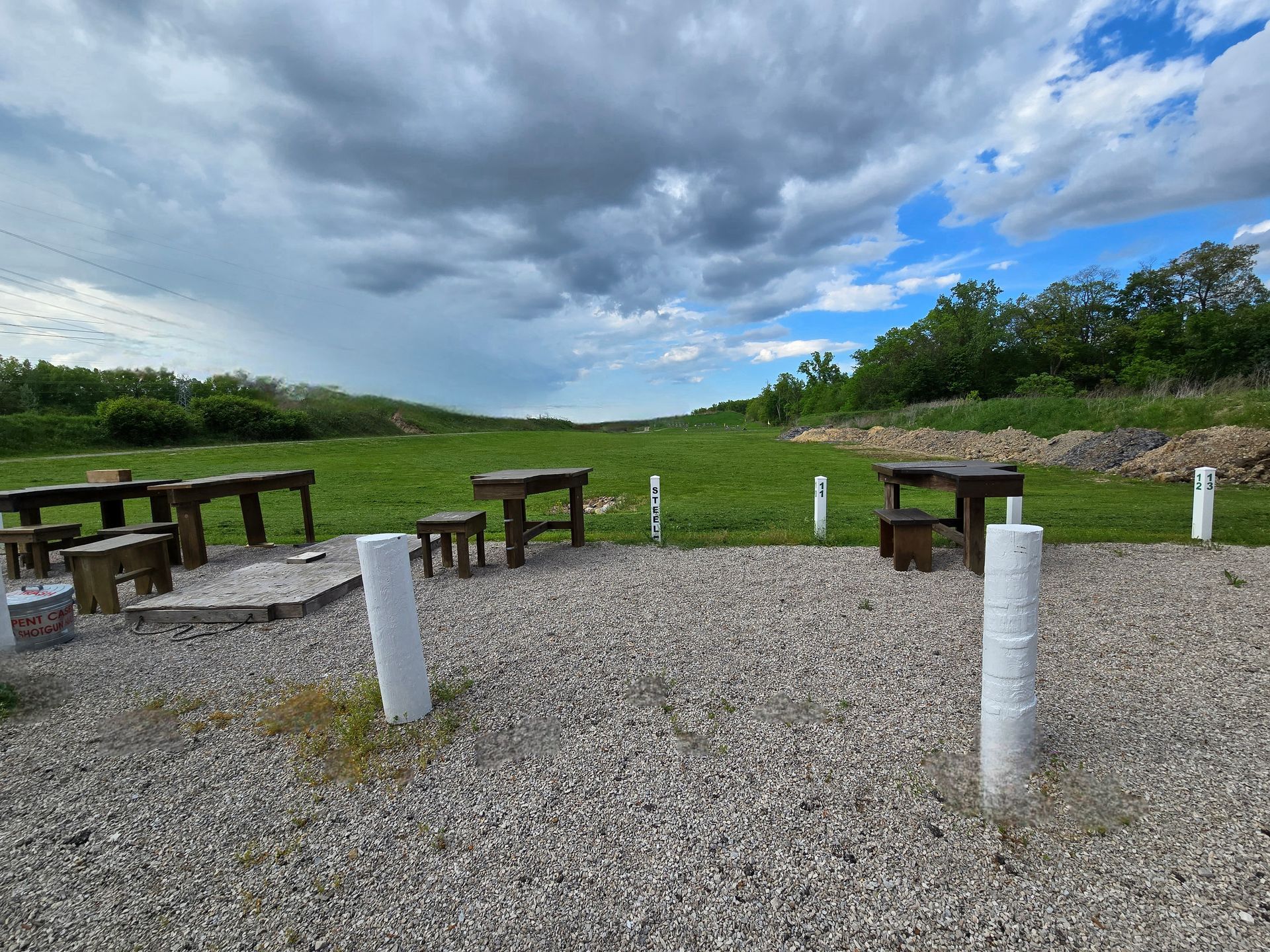 A graveled outdoor shooting range featuring several wooden benches and chairs under a cloudy sky.