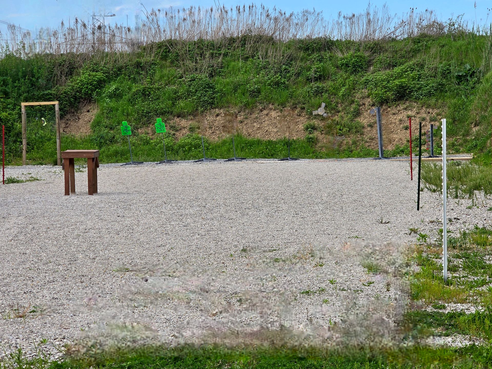 An outdoor gravel shooting range with target stands and green silhouettes set against a grassy earthen berm.