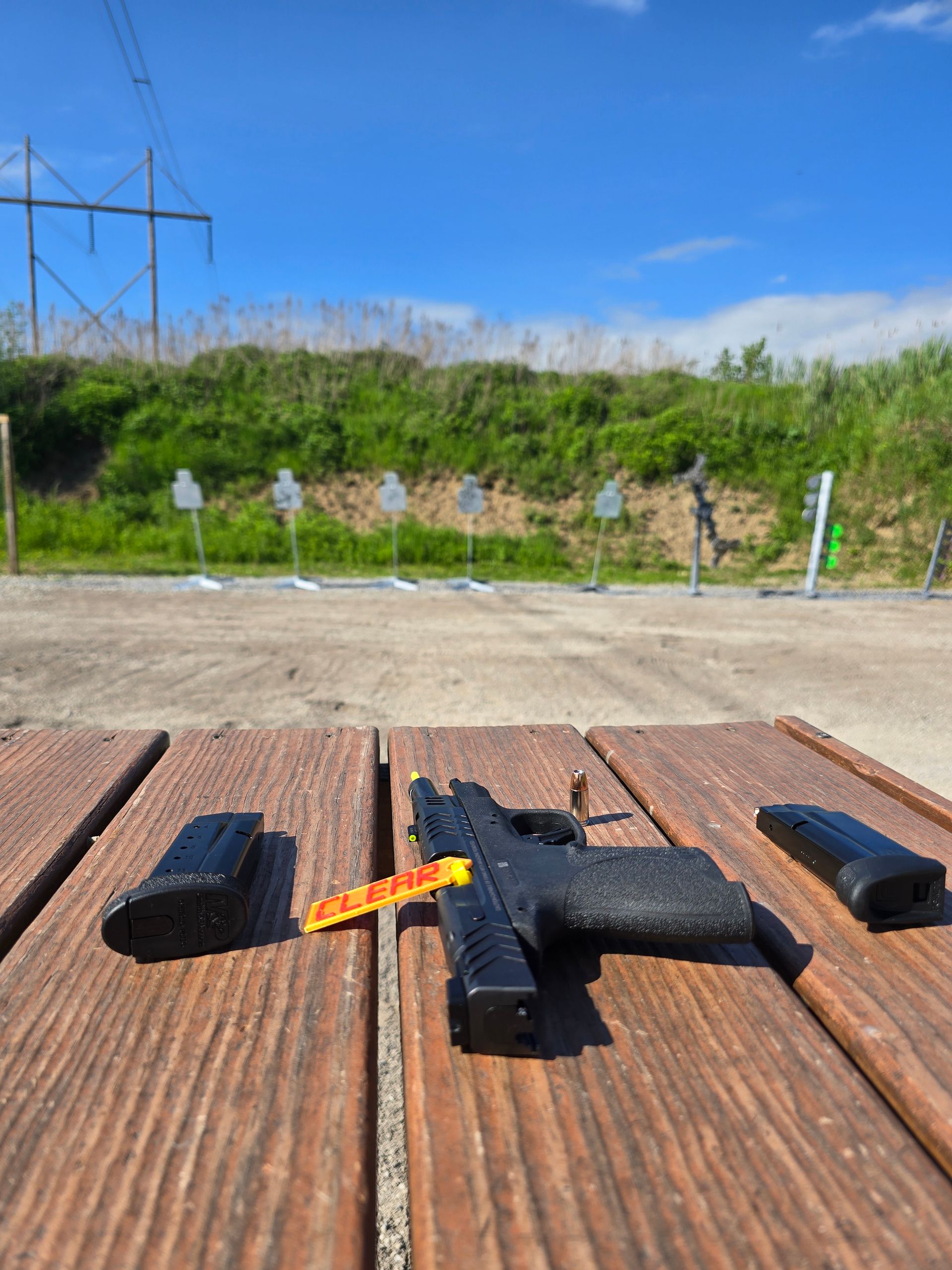 A black pistol with a yellow chamber flag sits on a wooden shooting bench with two magazines at an outdoor range.