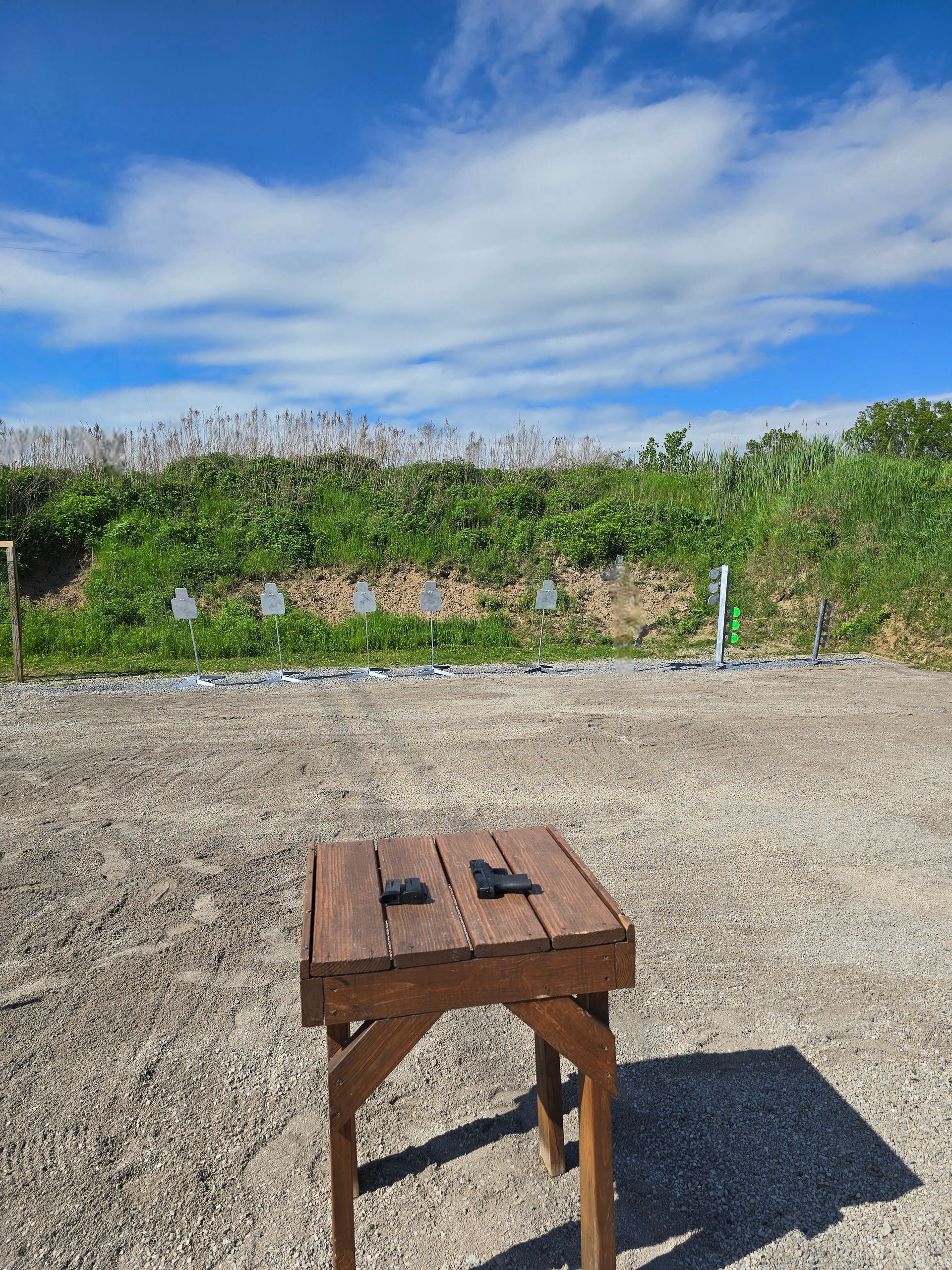 Two handguns rest on a wooden shooting bench at an outdoor range, facing a dirt berm with targets in the distance.