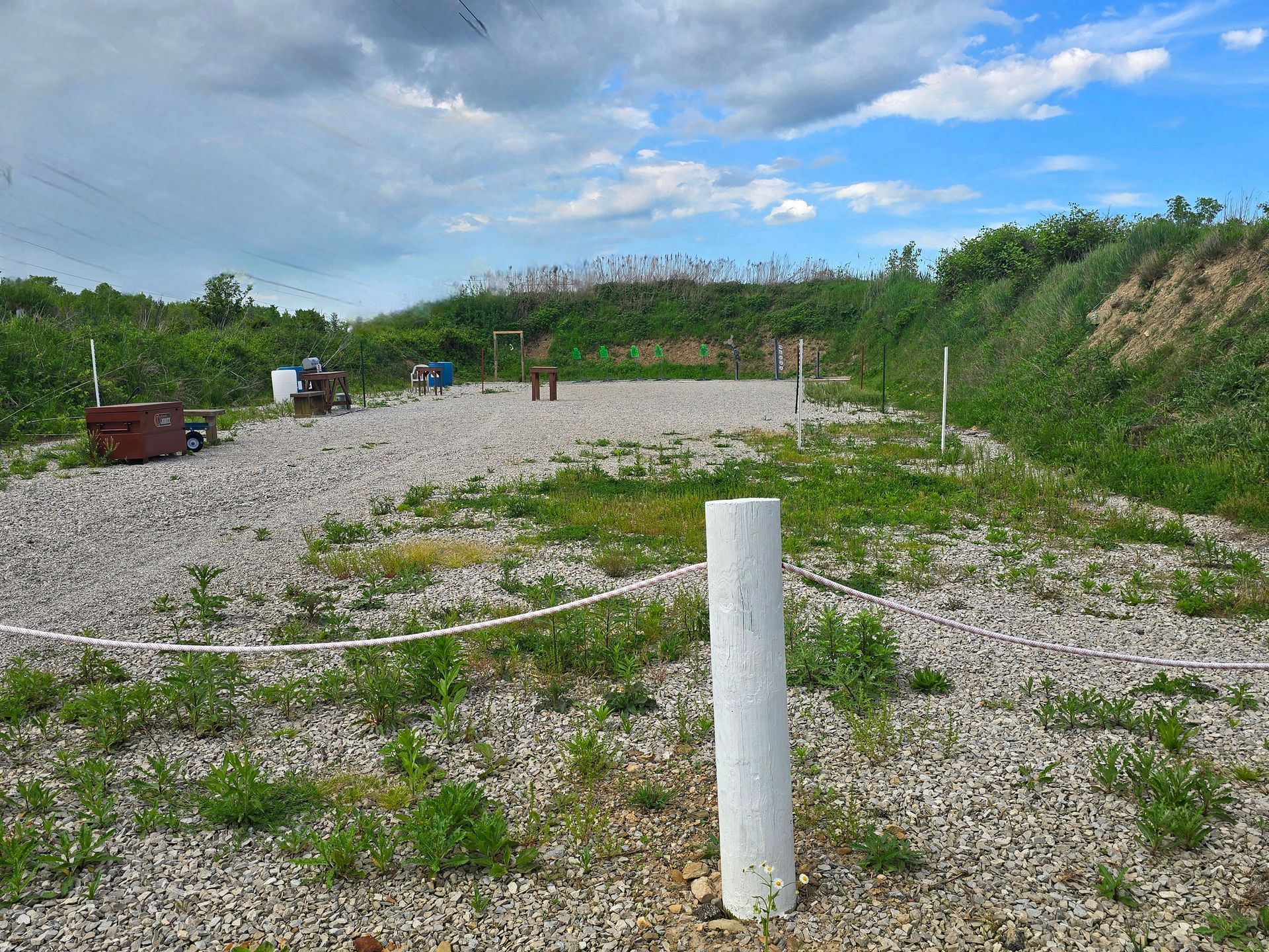 An outdoor shooting range featuring a gravel ground, target stands, and a protective earthen berm under a cloudy sky.