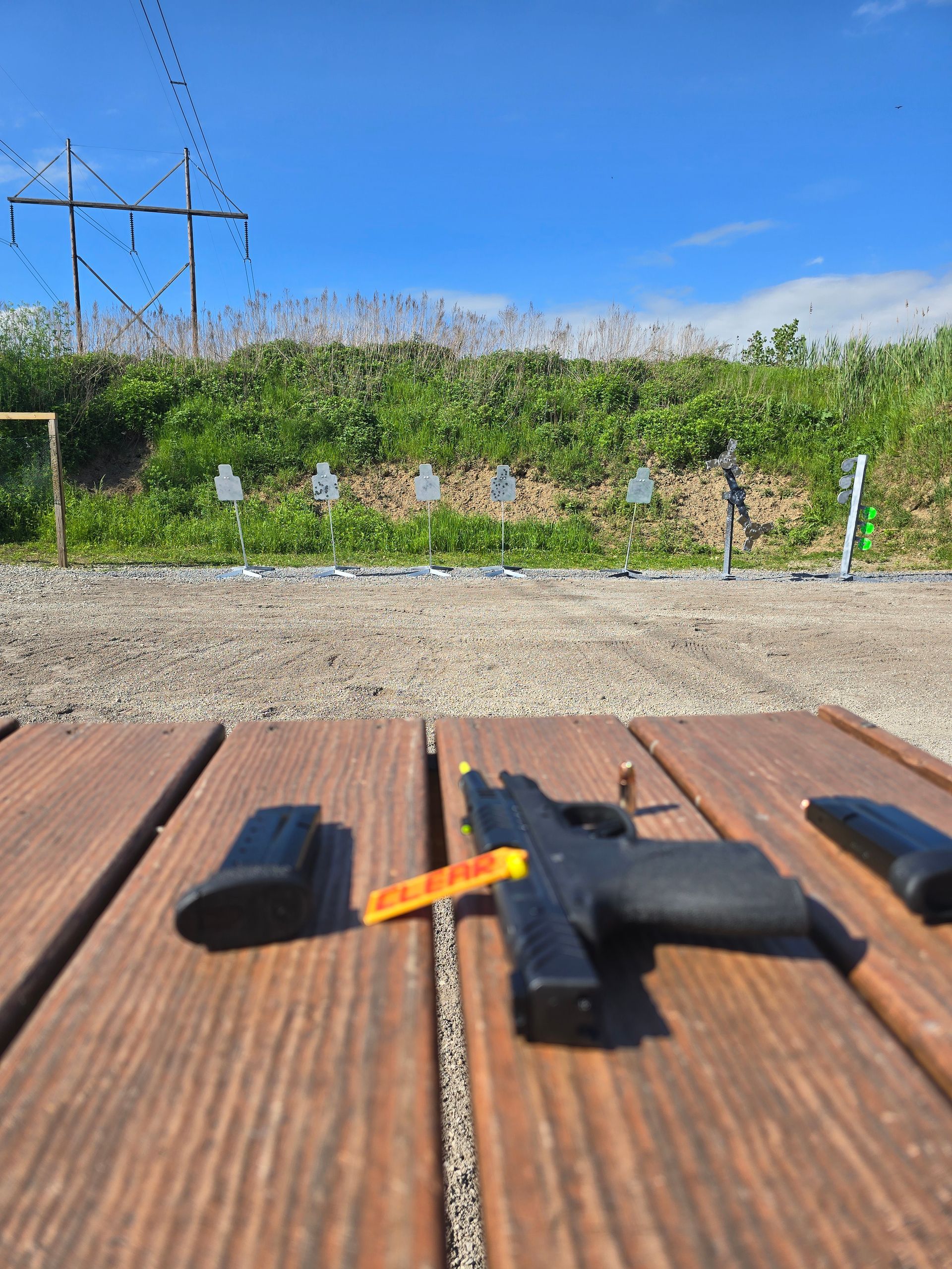 A handgun with a yellow safety flag, two magazines, and a bullet sit on a wooden table at an outdoor shooting range.