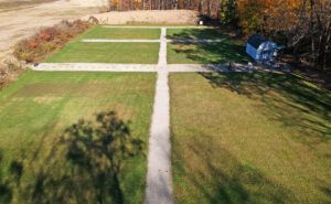 Aerial view of a grass field with a central gravel path intersecting two horizontal paths, with a shed in the corner.