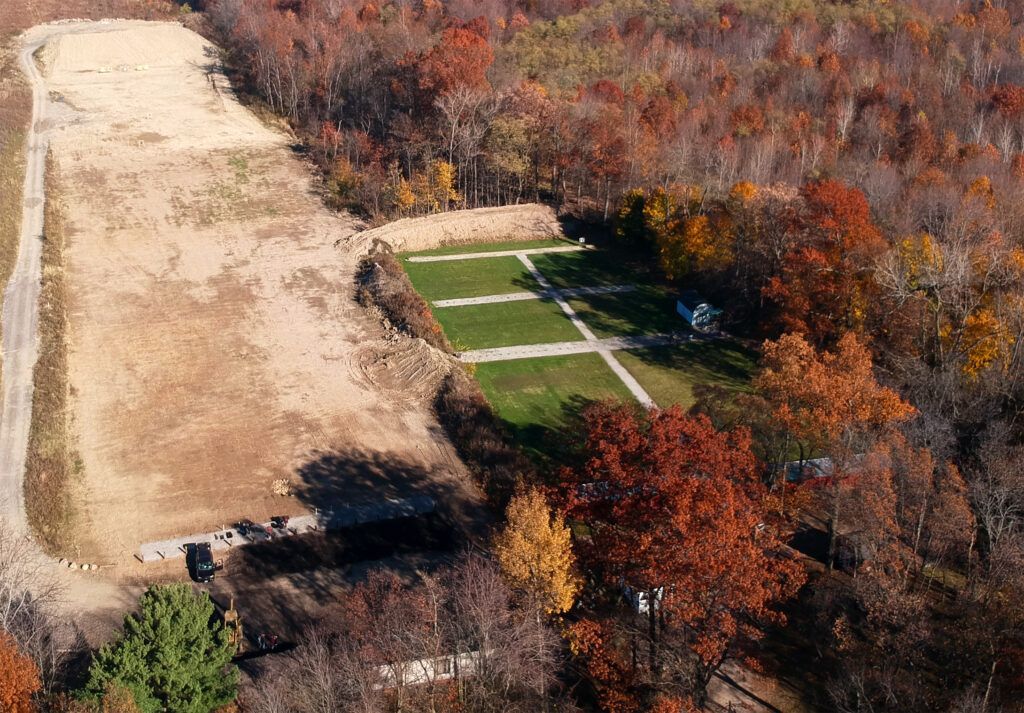 Aerial view of a rectangular outdoor shooting range carved into a wooded, autumnal landscape with a cleared dirt area.