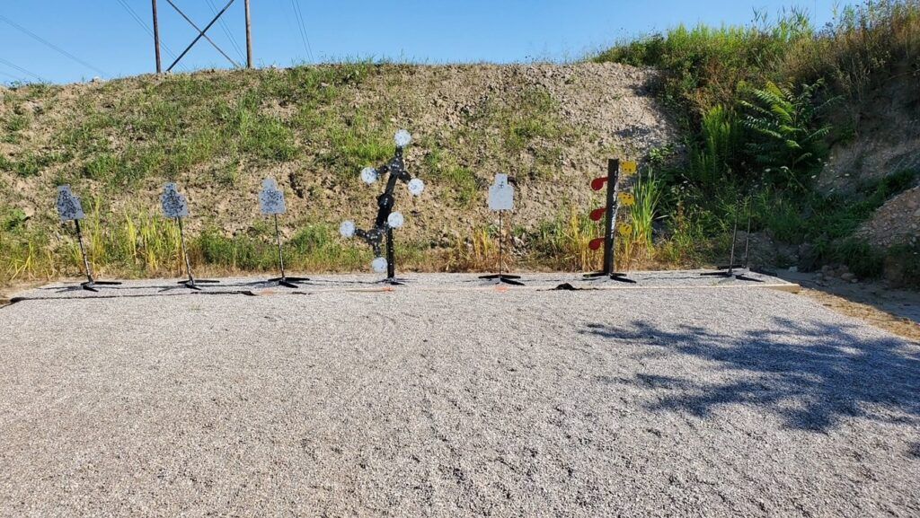 Metal target stands with circular plates arranged on a gravel shooting range against a grassy earthen berm.