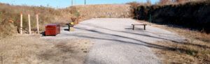 An outdoor shooting range featuring a gravel ground, target stands, a red shooting bench, and a wooden bench.