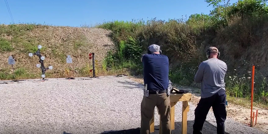 Two people shooting at metal targets at an outdoor range with a dirt berm in the background.