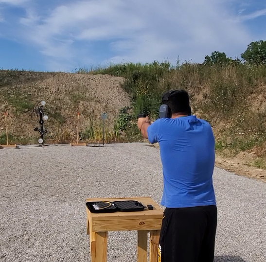 A person in a blue shirt firing a handgun at targets on a gravel outdoor shooting range.