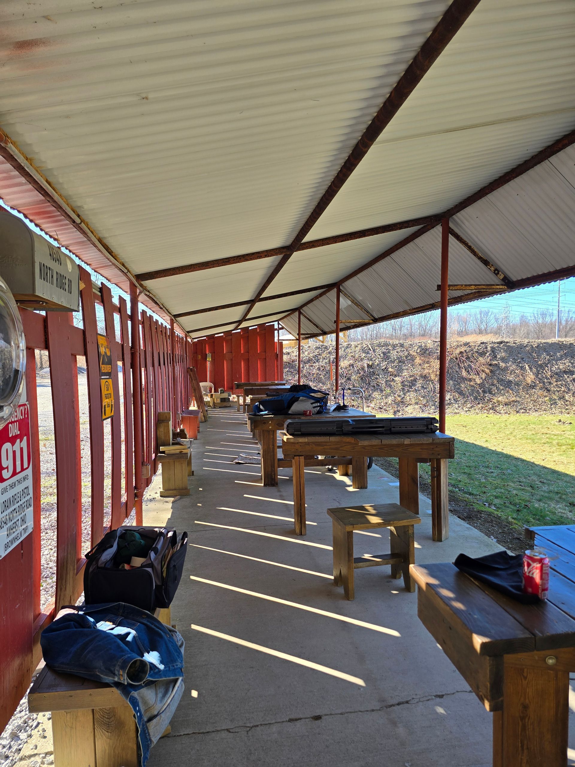 An outdoor shooting range with covered wooden benches and stools arranged in a row facing a dirt berm.