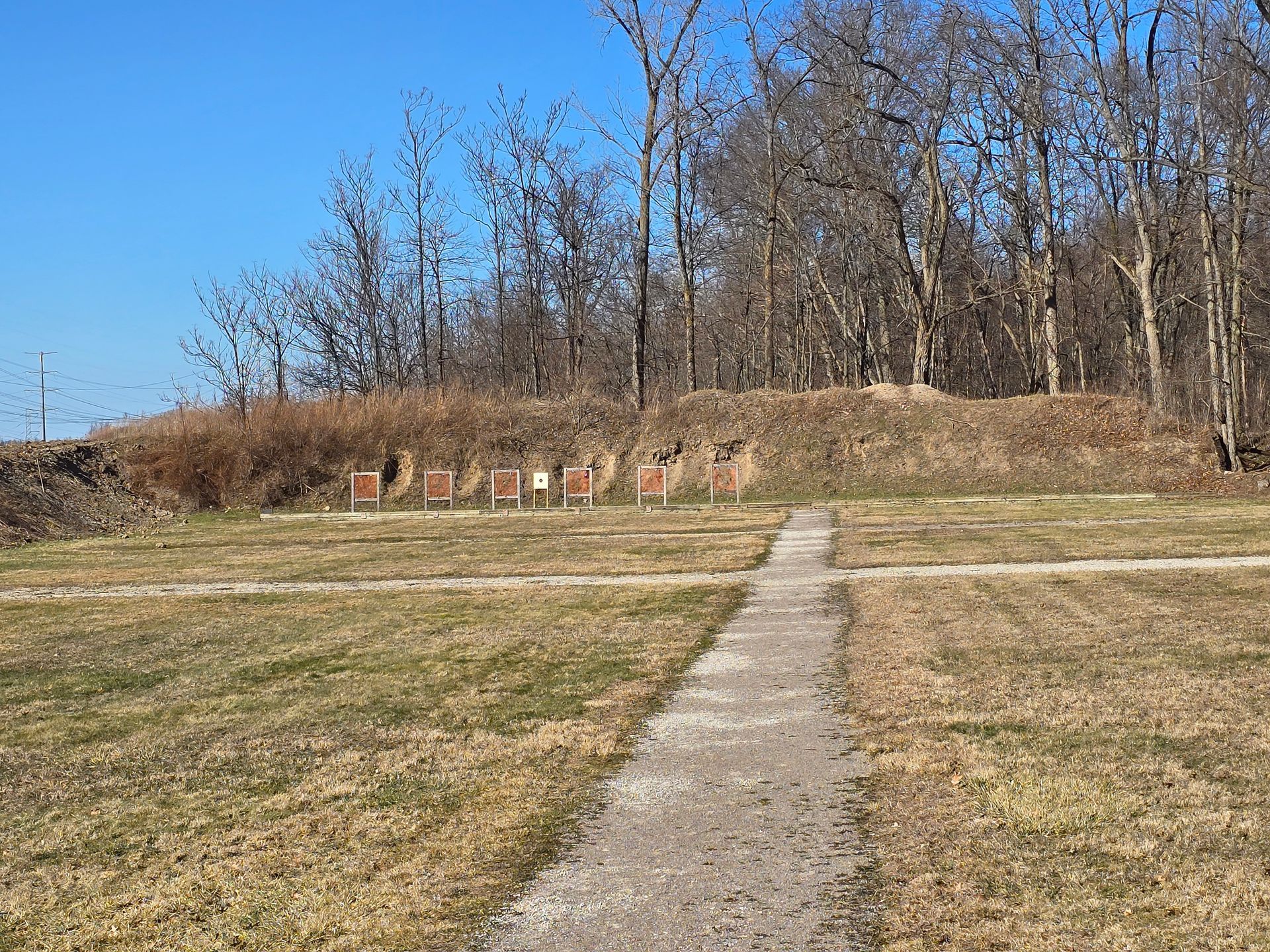 An outdoor shooting range with target stands set against a grassy earthen berm and a background of bare trees.