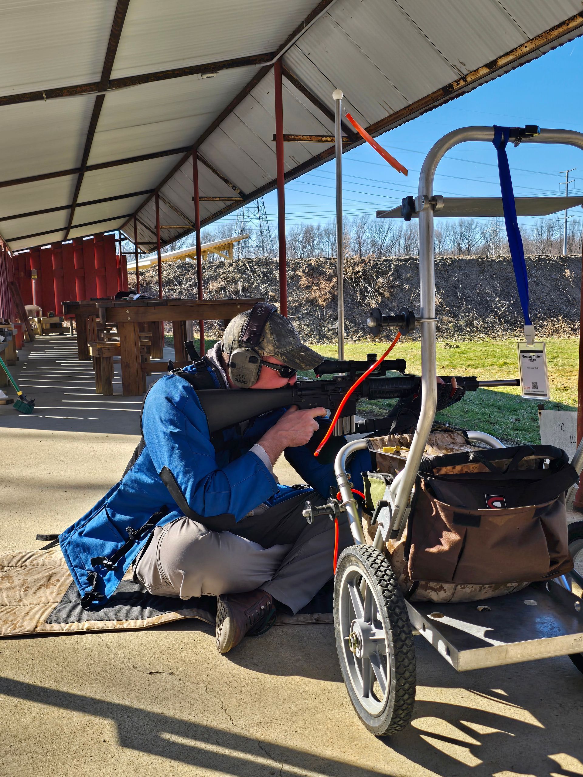 A person sits on a shooting range mat, using a rifle supported by a specialized cart under a covered shelter.