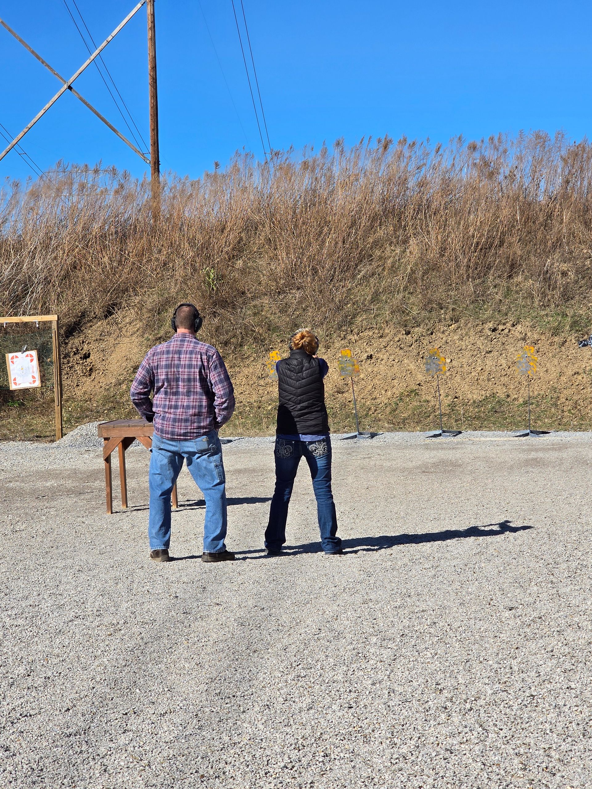 Two people stand side-by-side at a gravel outdoor shooting range, facing targets placed against a dirt berm.