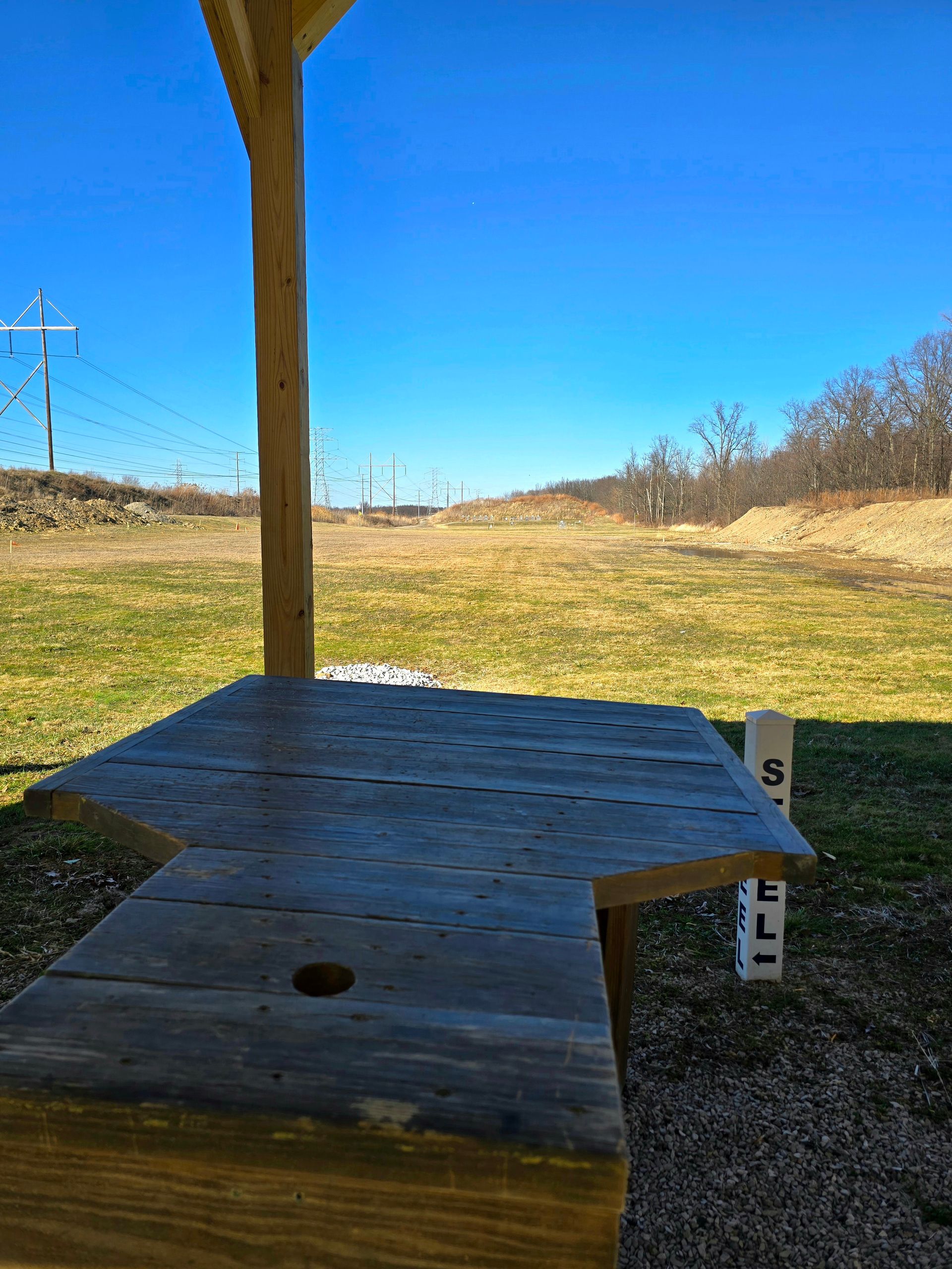 A wooden shooting bench sits under a pavilion at an outdoor range, facing a grassy field under a bright blue sky.