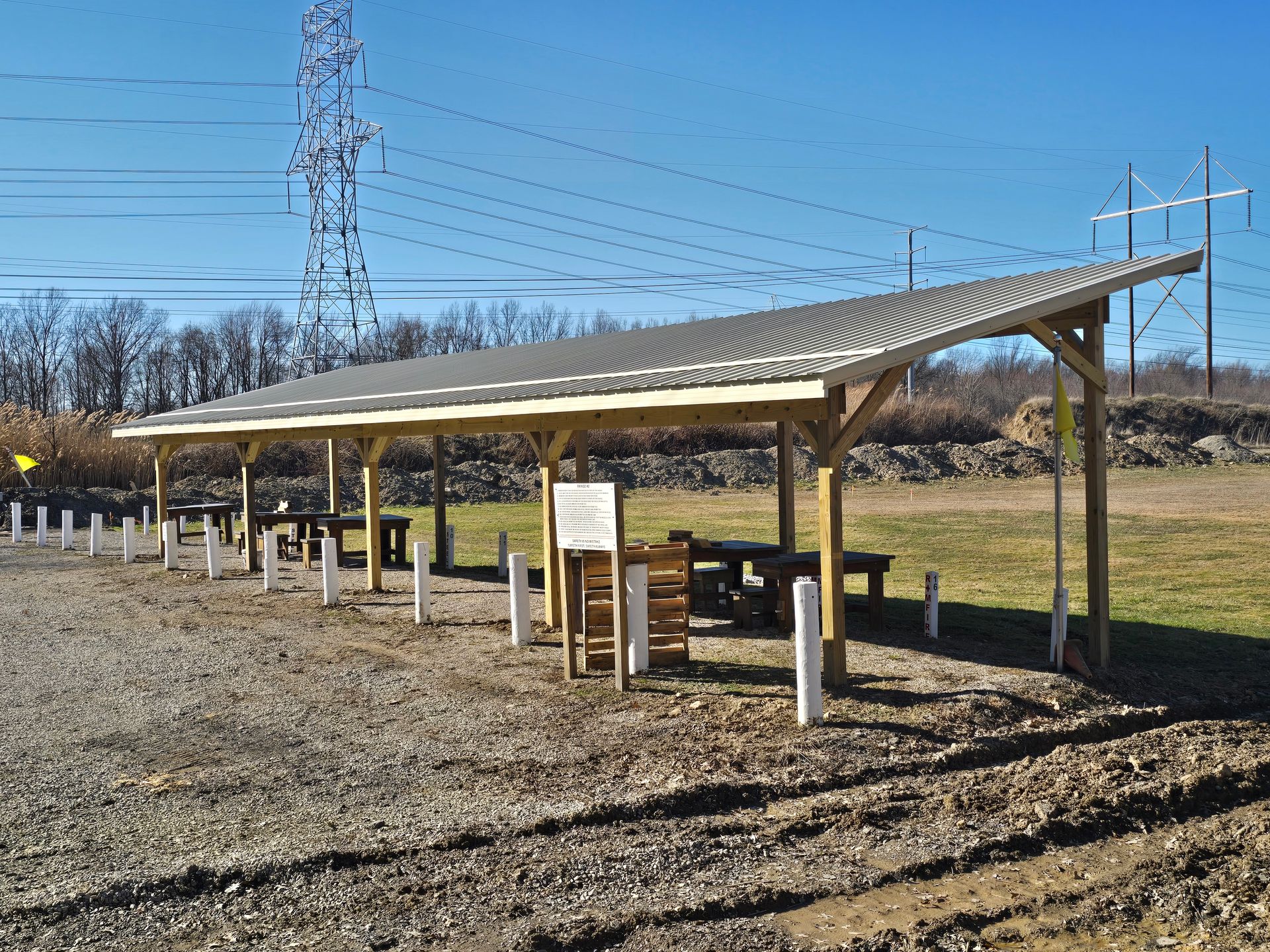 A long wooden shelter with a sloped metal roof stands over benches on a gravel lot under a clear blue sky.