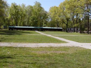A long, low building with a green roof sits in a grassy park clearing with walking paths and tall trees.