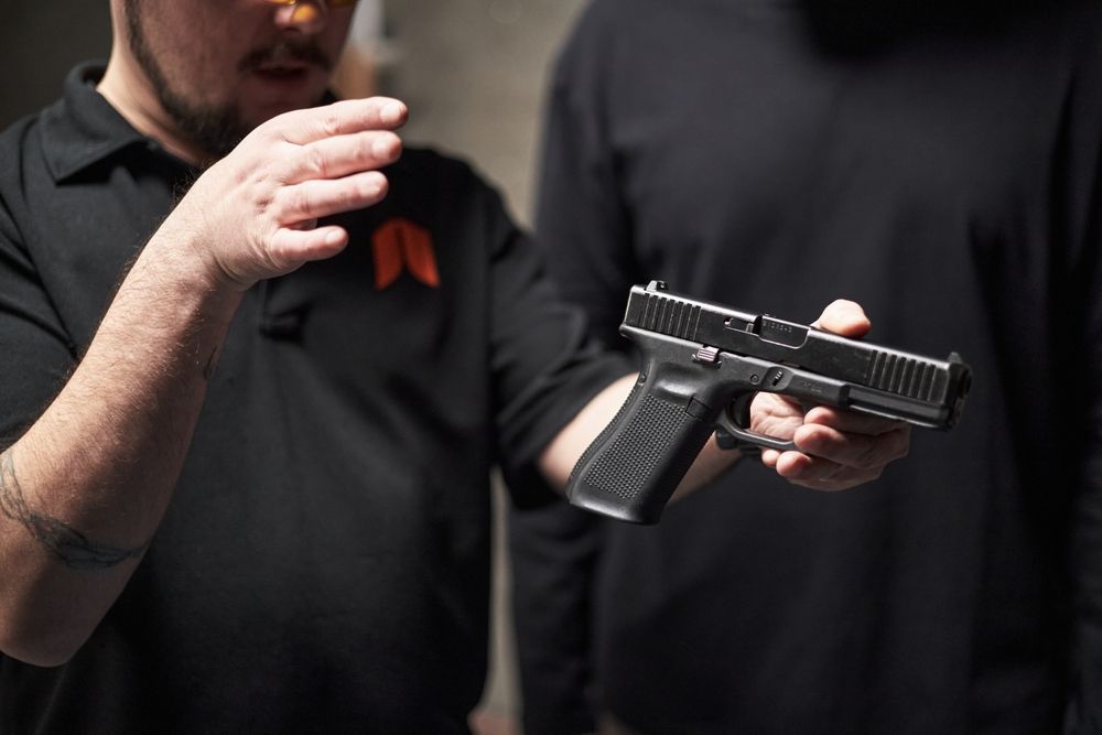 A person in a black shirt gestures while another person holds a black semi-automatic pistol in an indoor setting.