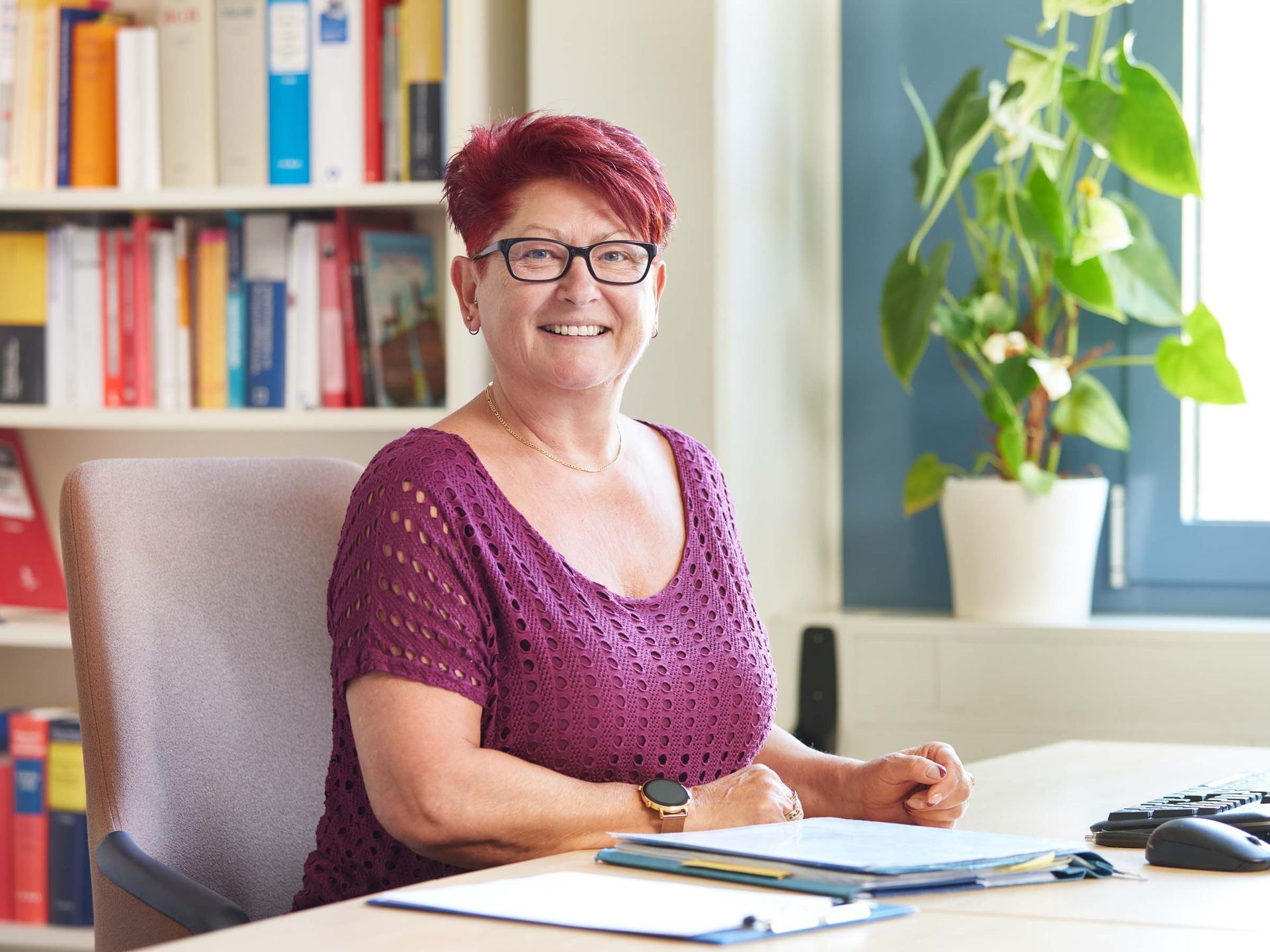 A woman is sitting at a desk wearing glasses and a purple shirt.