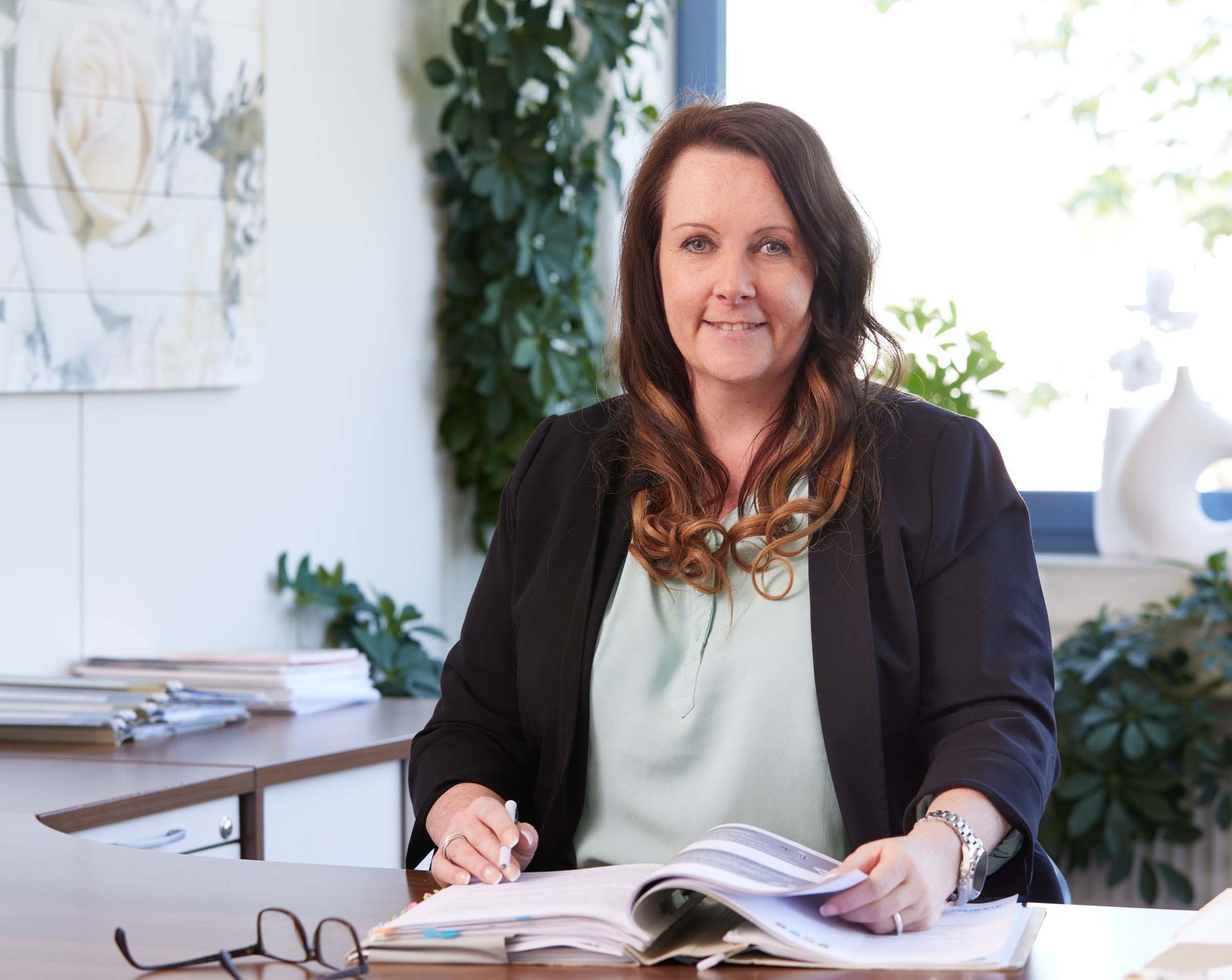 A woman is sitting at a desk reading a book and smiling.