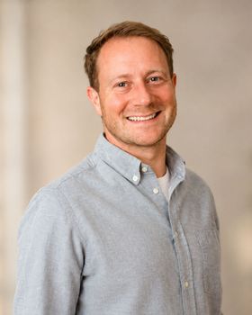 A man in a grey shirt is smiling in front of a black background.
