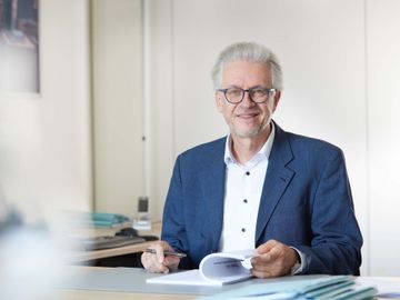 A man in a suit and glasses is sitting at a desk holding a book.