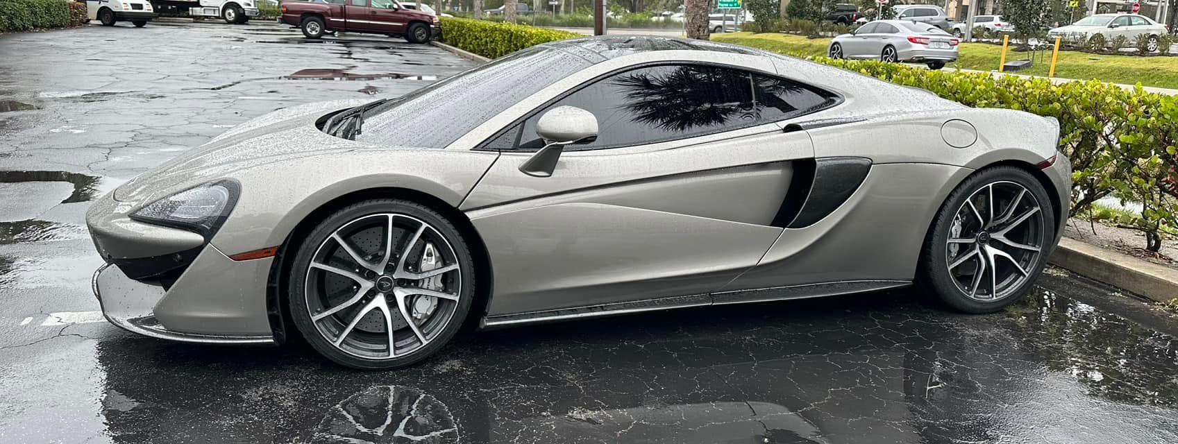 Silver McLaren sports coupe with dark side glass, shown in profile on wet pavement.