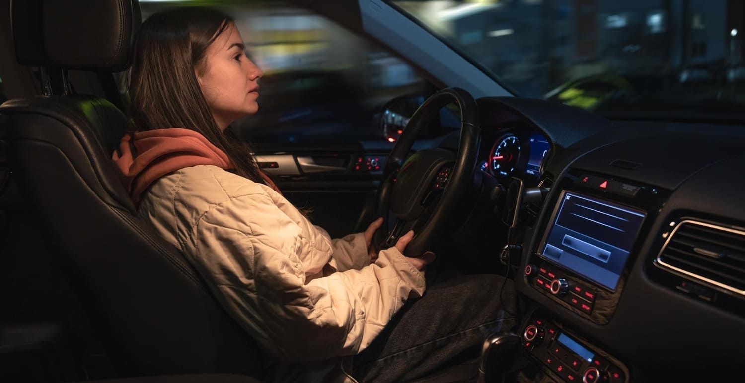 Driver seated at the wheel of a car at night with the dashboard illuminated.