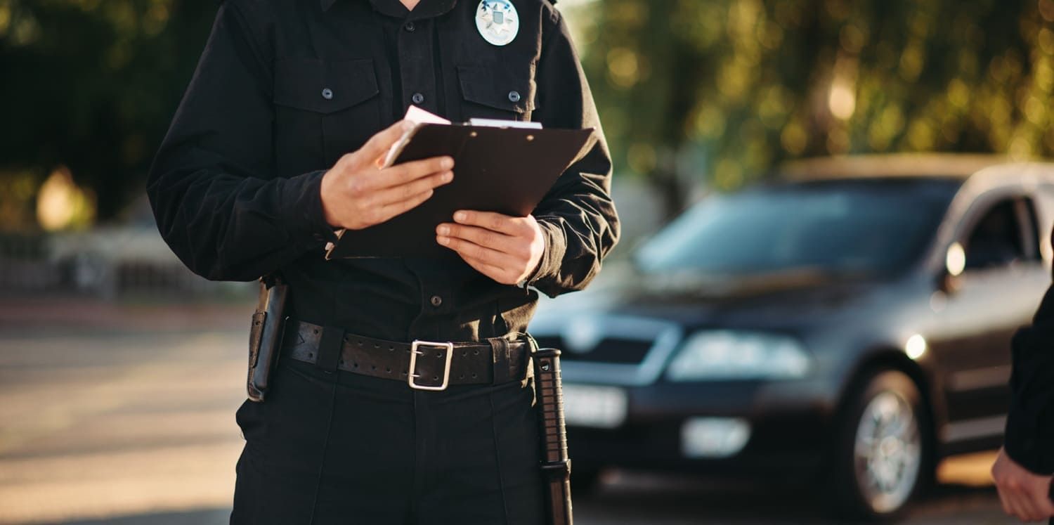 Uniformed officer holding a clipboard beside a parked car.
