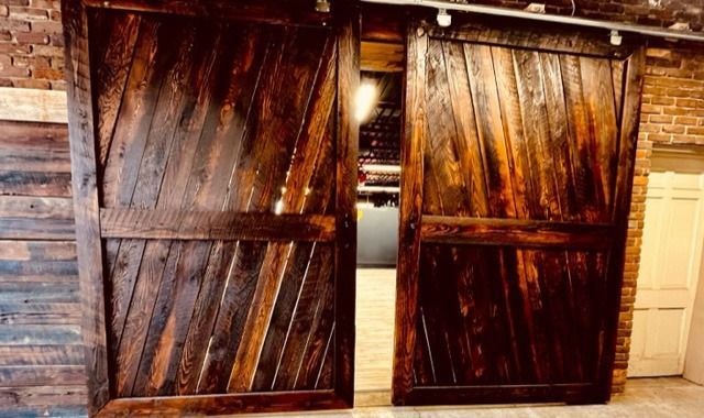Rustic wooden barn doors partially open, revealing a dimly lit interior against a brick wall.