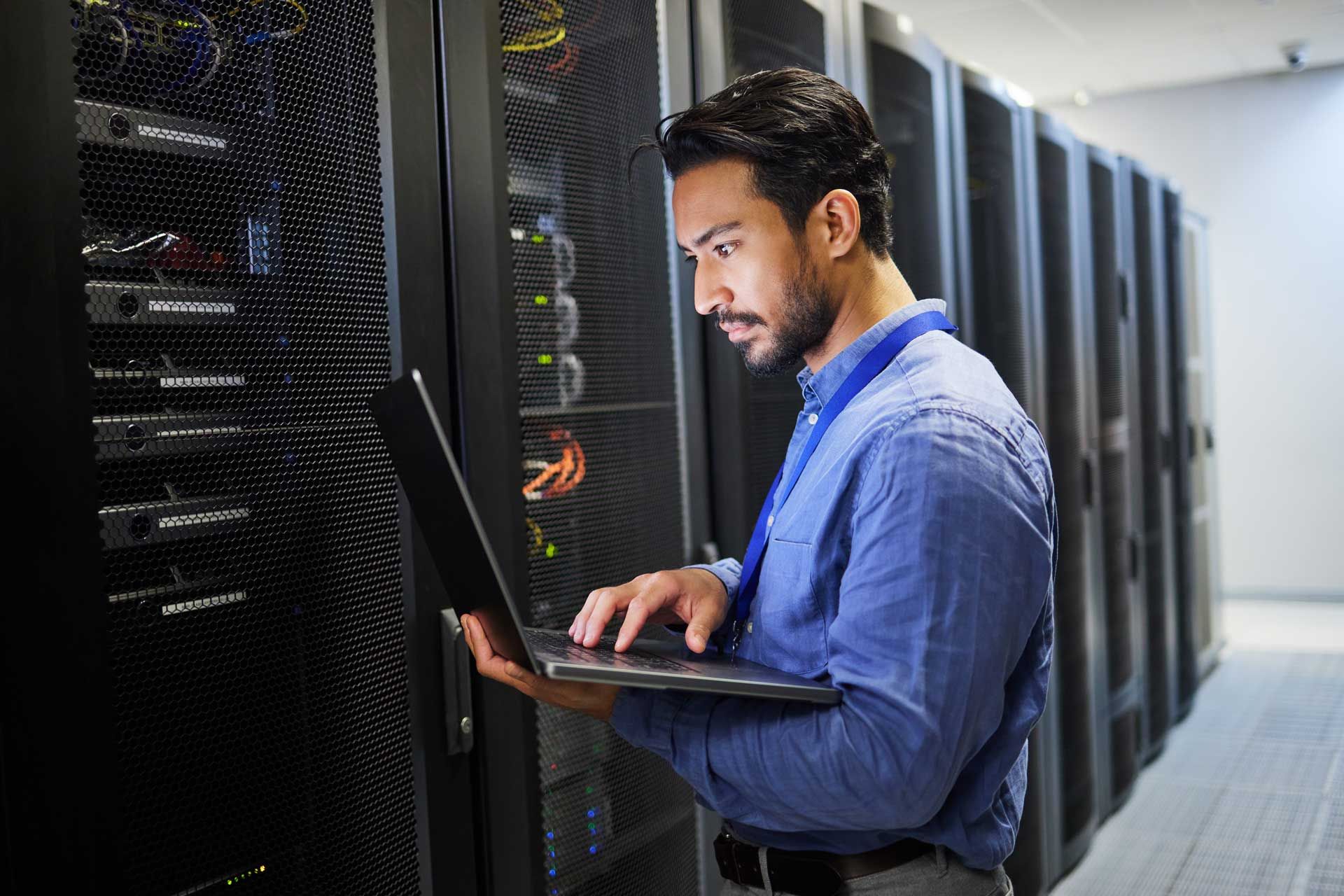 A man is using a laptop in a server room.