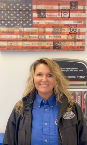 A woman in a blue shirt and black jacket is standing in front of an american flag.