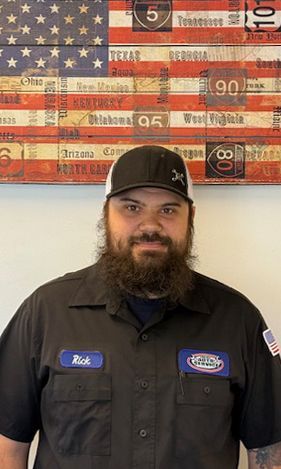 Man in black work shirt and cap, standing in front of an American flag made of license plates.