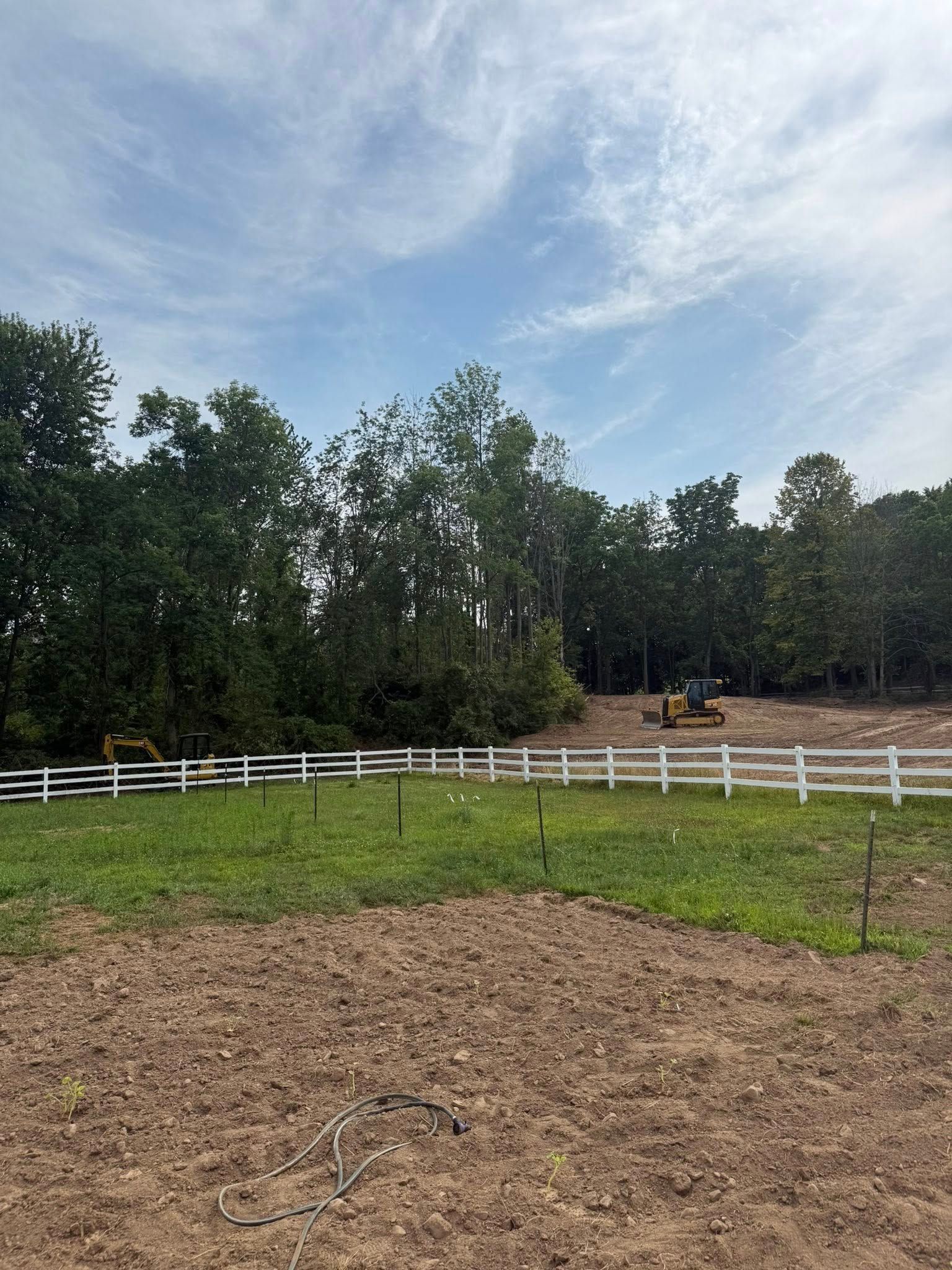 A field with a white fence, trees in the background, and construction equipment. Cloudy blue sky above.