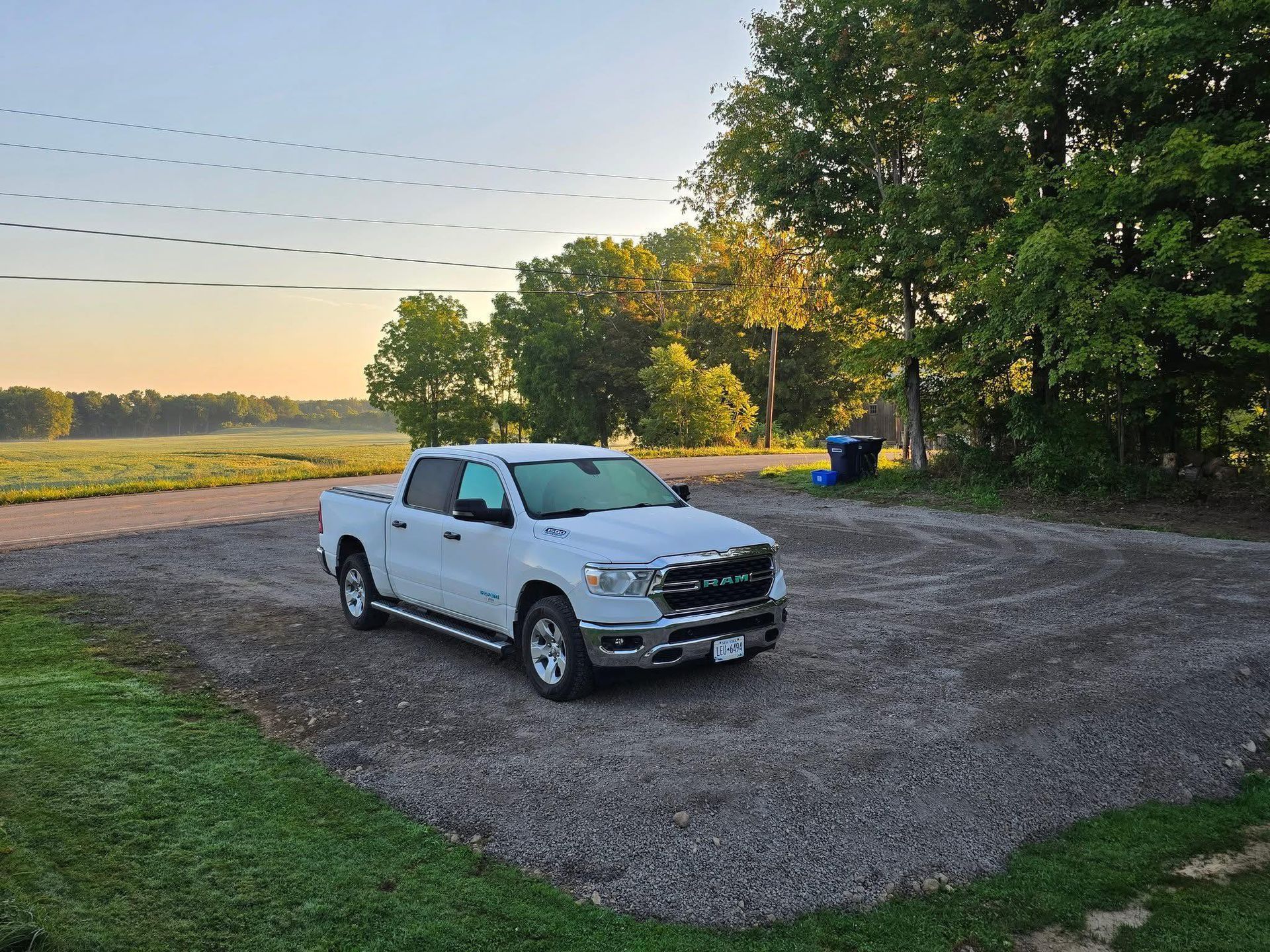 White pickup truck parked on gravel, trees and field in the background. Blue sky, morning light.