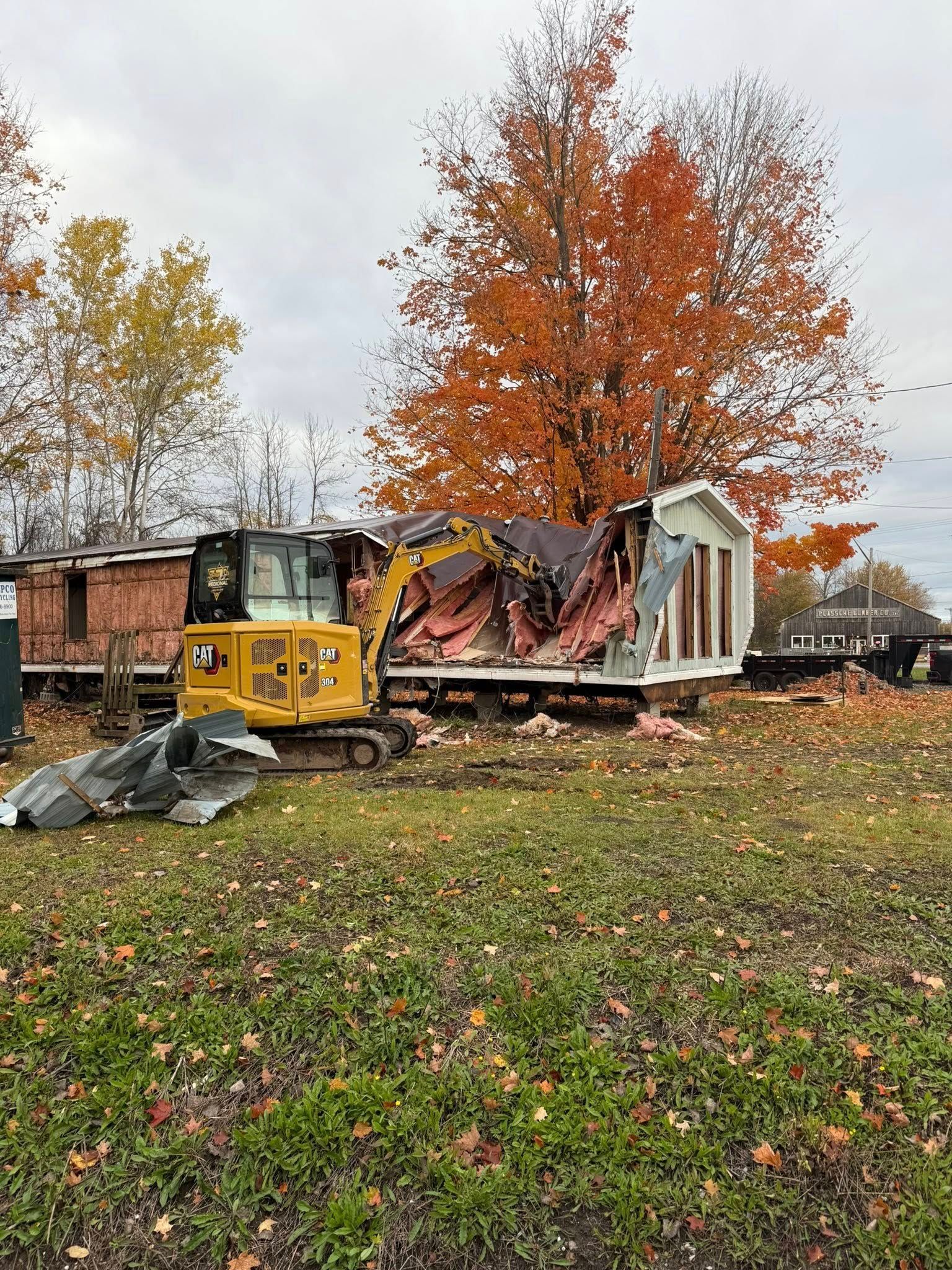 Excavator demolishing a white mobile home on a grassy lot, fall foliage in the background.
