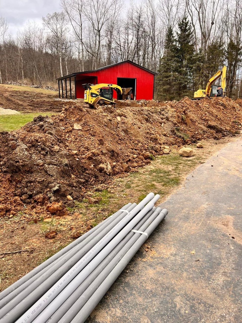 Construction site with red barn, excavators, and gray pipes. Earth mound in front.