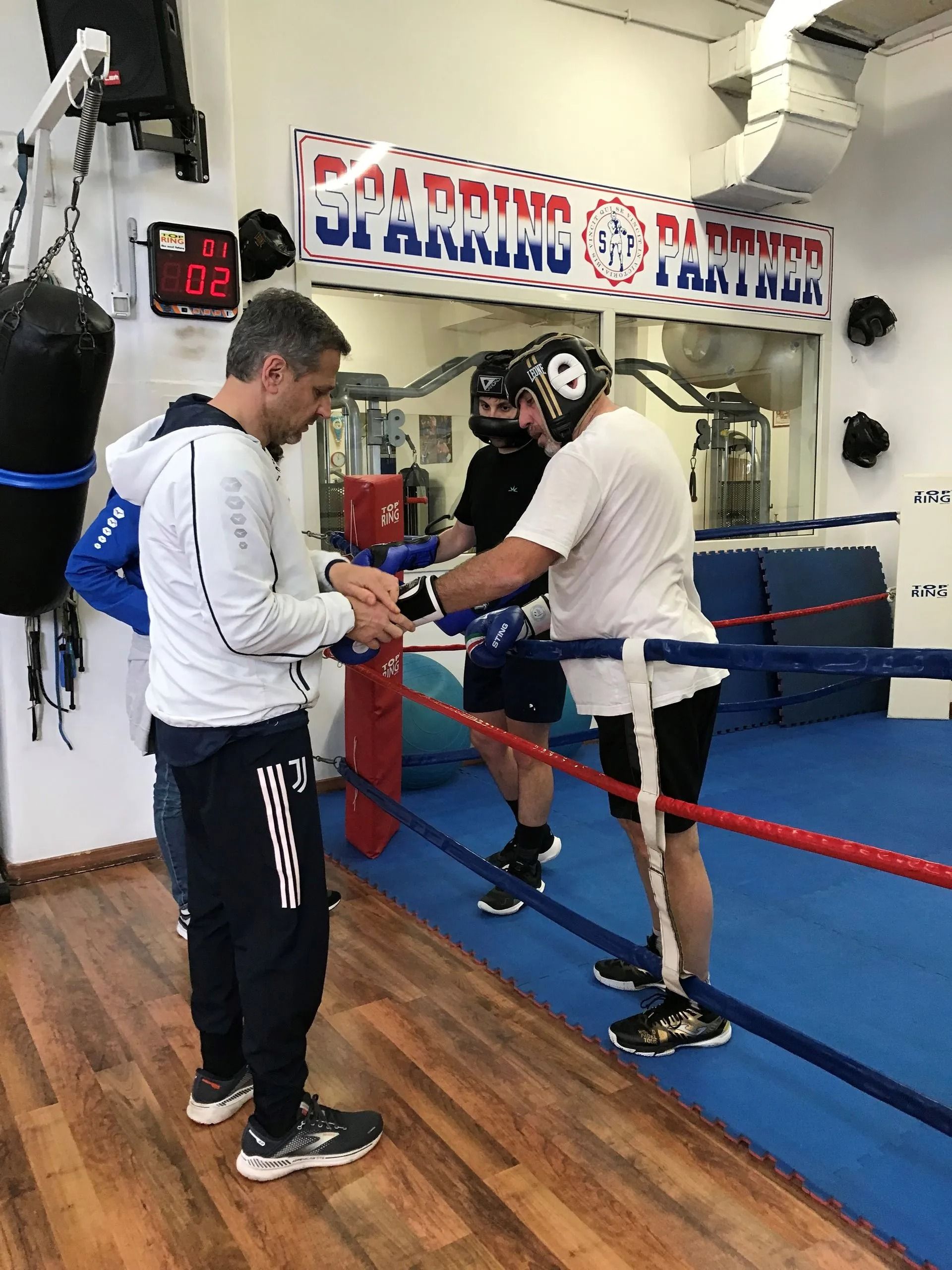 Due pugili si allenano in palestra, indossando caschetto e guantoni, con un allenatore che li osserva da vicino.