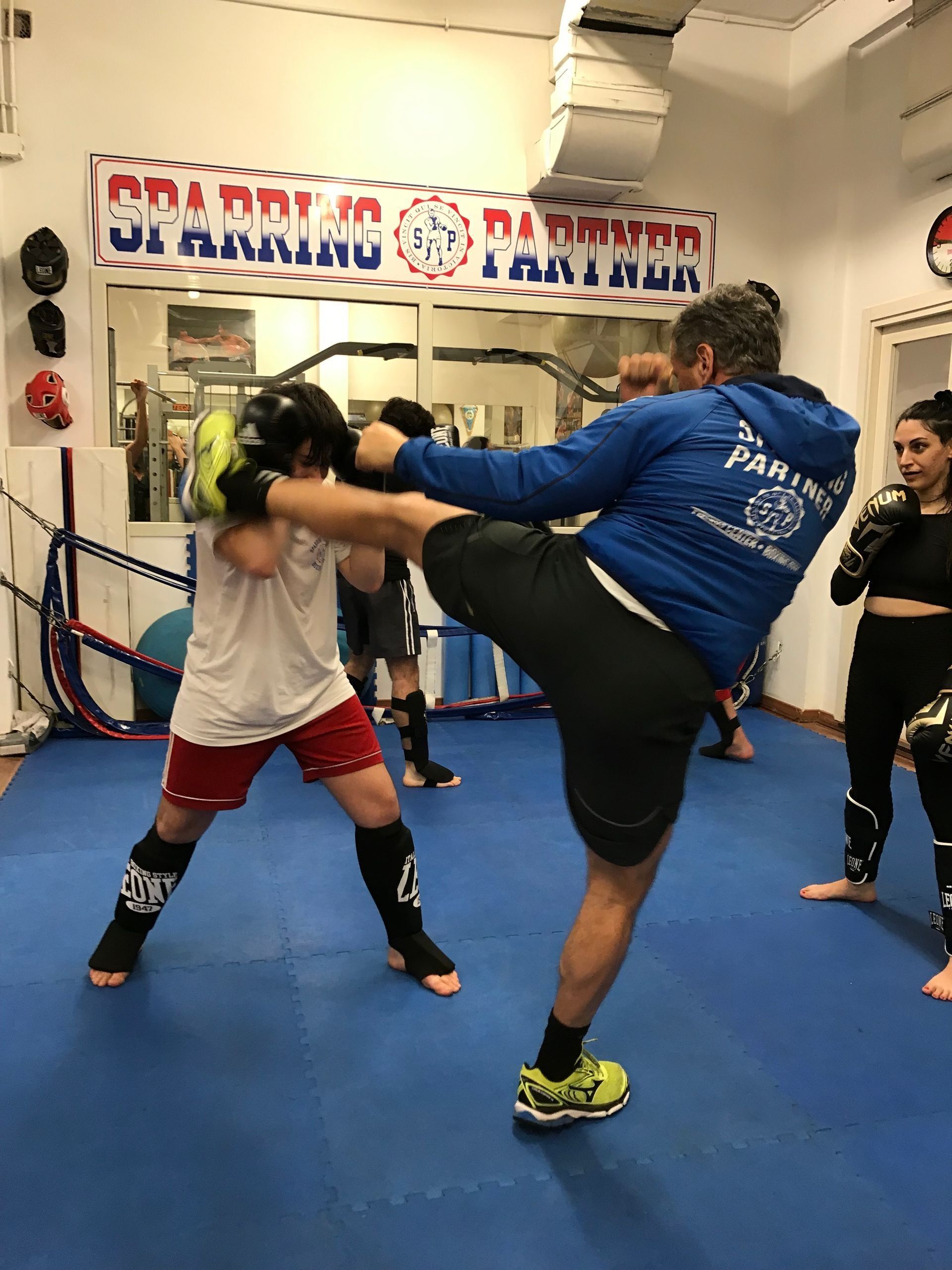 Due persone si allenano in una palestra di arti marziali, una delle due sferra un calcio alto su dei tappetini blu.