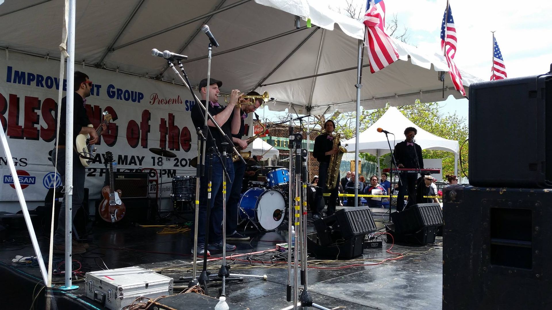 Band performing on an outdoor stage under a canopy, with drums, guitars, flags, and speakers in view.